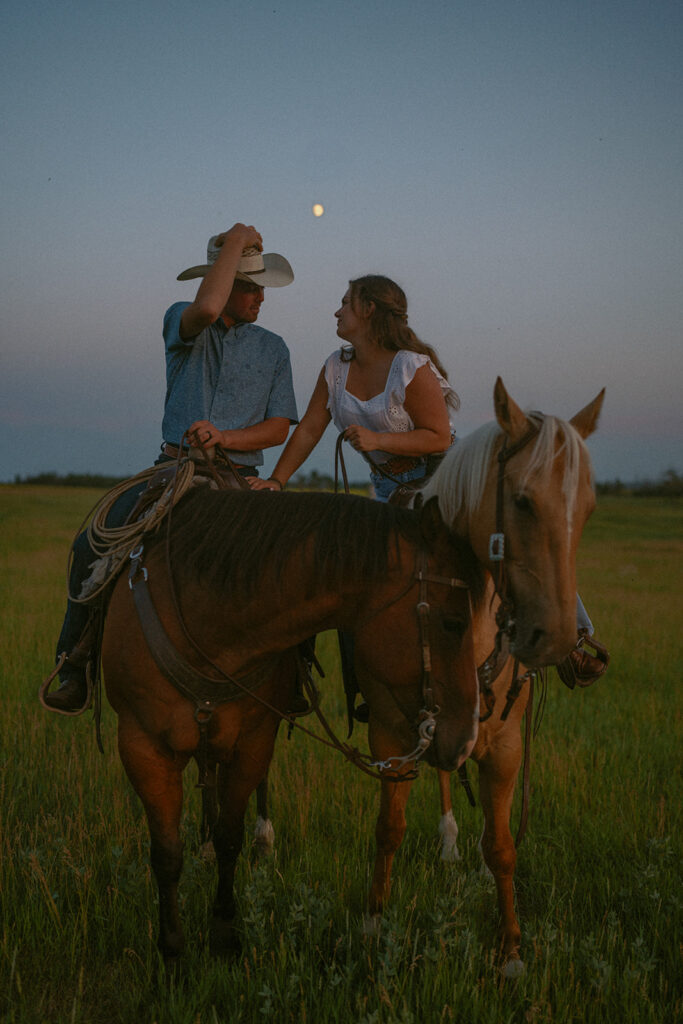 couple leaning into each other for a kiss while both on horses at blue hour.