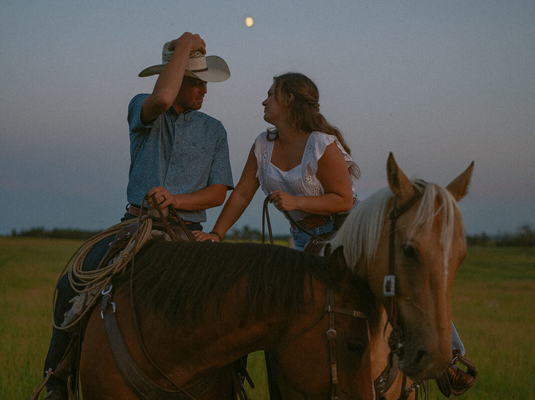 couple leaning into each other for a kiss while both on horses at blue hour.