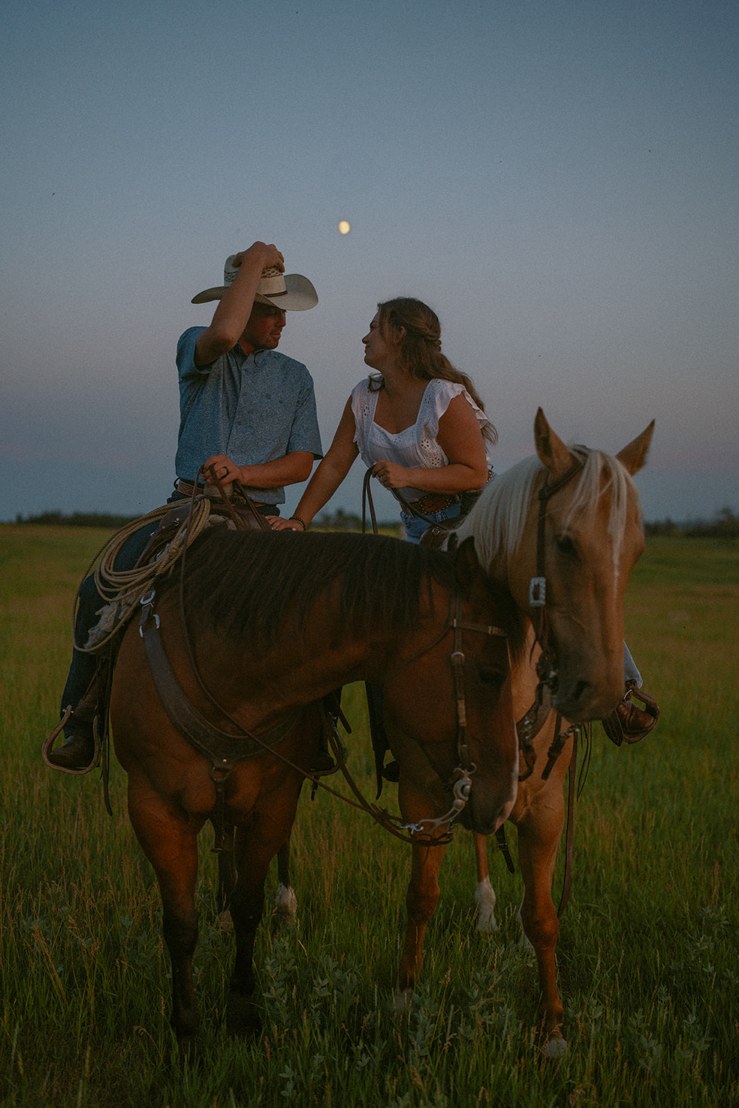 couple leaning into each other for a kiss while both on horses at blue hour.